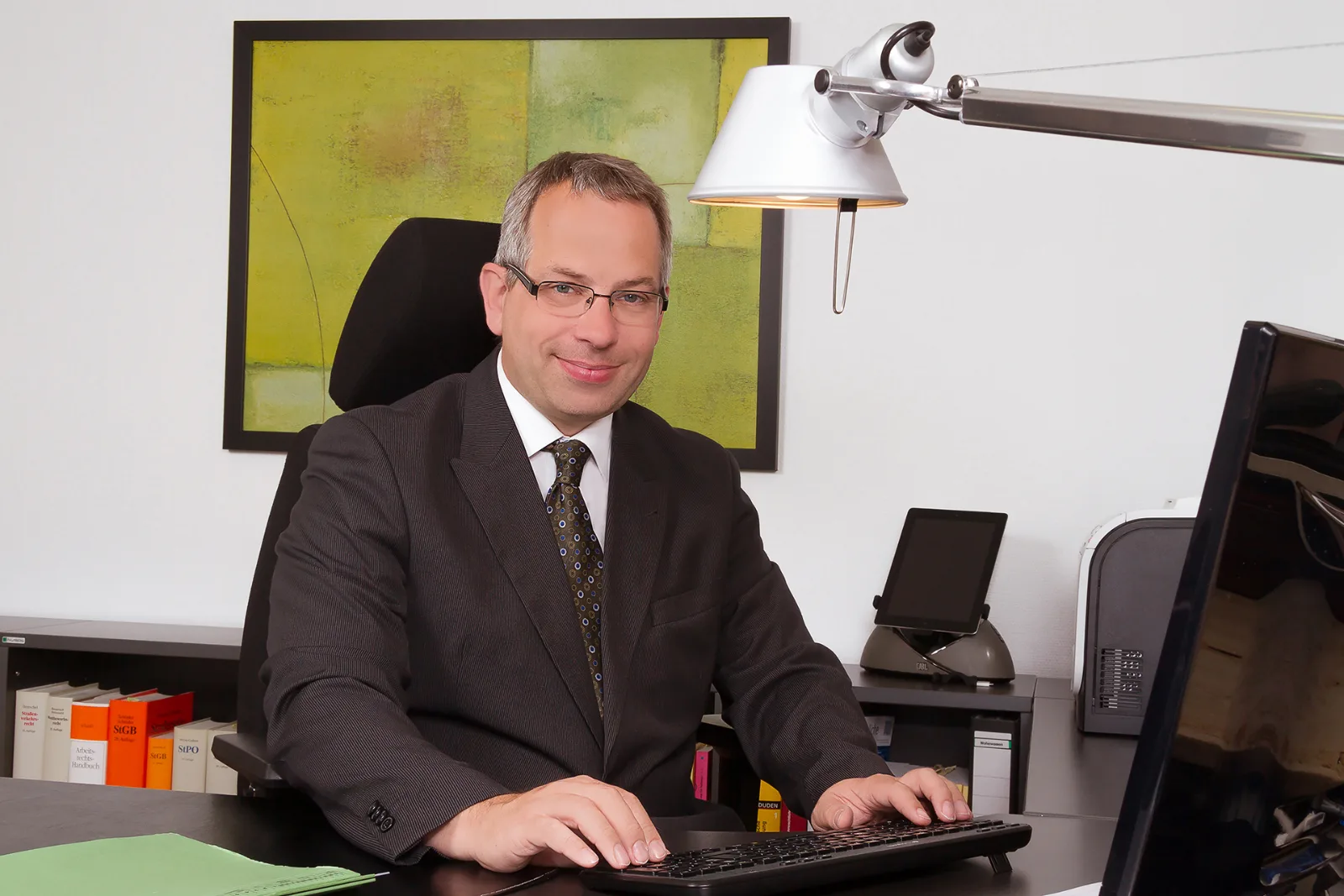 Geschäftsmann im Anzug am Schreibtisch, im Hintergrund Aktenrücken und ein modernes Wandbild