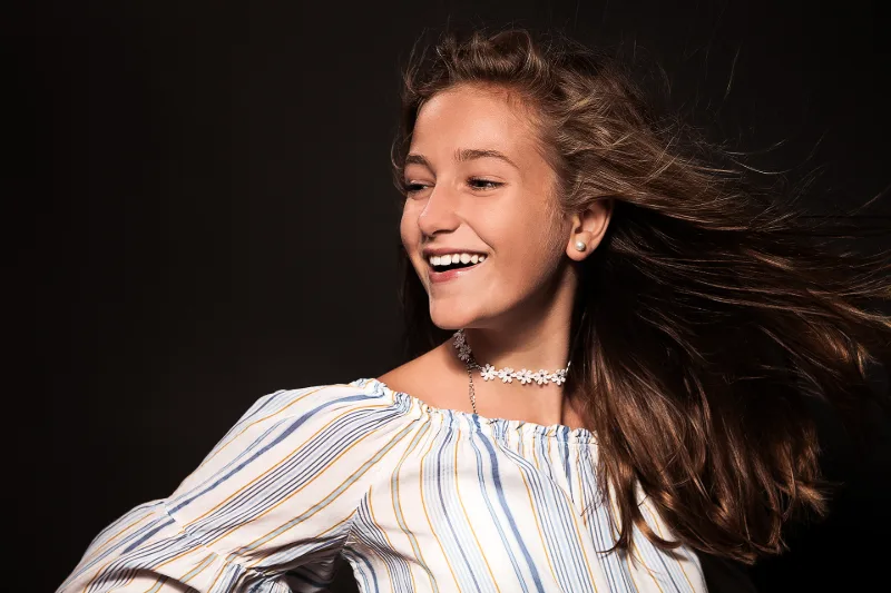 A young woman with wind-blown hair, laughing as she looks to the side, against a black background