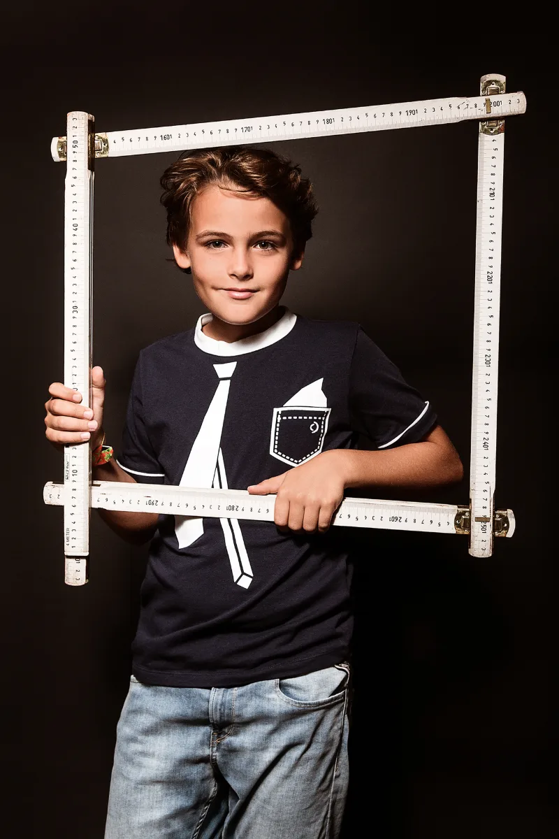 A boy framing his face with a folding carpenter's rule bent into a square against a black background