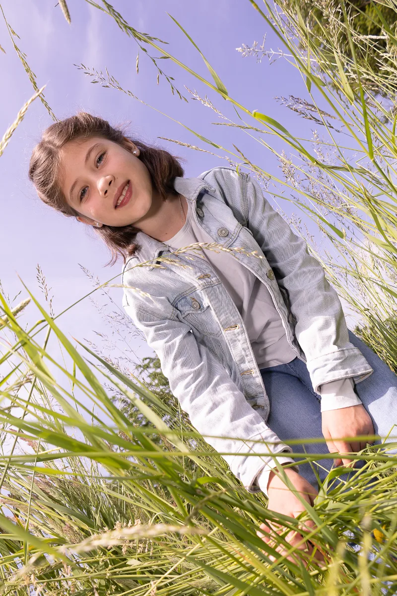 A girl in a denim jacket sitting in tall grass, shot from below against a lilac sky
