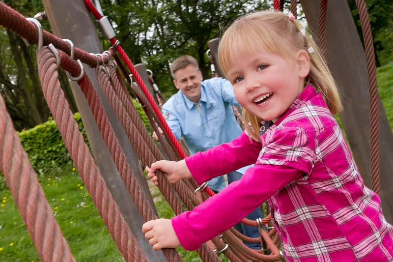 A girl in a pink shirt laughing as she climbs a rope net, her father watching behind