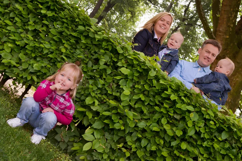 A little girl with a finger to her lips at a tall hedge, her parents with two toddlers behind it