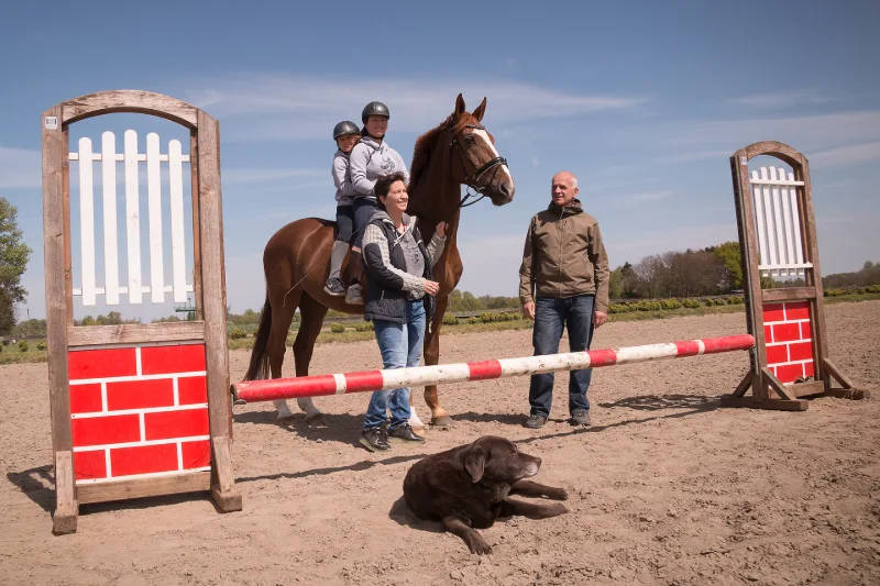 A family on the riding arena with a chestnut horse and dog in front of a red-and-white show jump