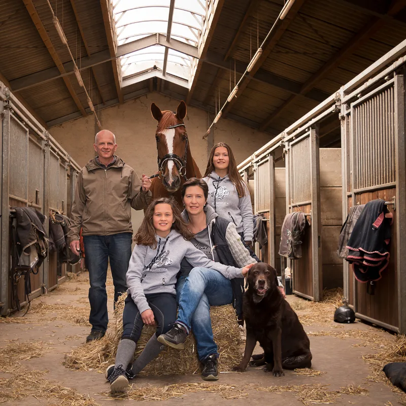 A family with a chestnut horse and brown Labrador in the stable aisle, the daughters in equestrian hoodies