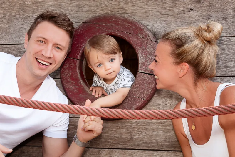 A toddler peeking through the round porthole of a play ship, parents laughing on either side
