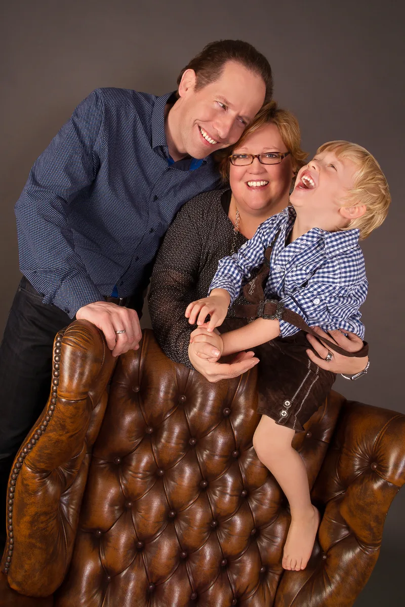 Parents with their laughing son in traditional dress on a brown Chesterfield chair against a grey studio backdrop
