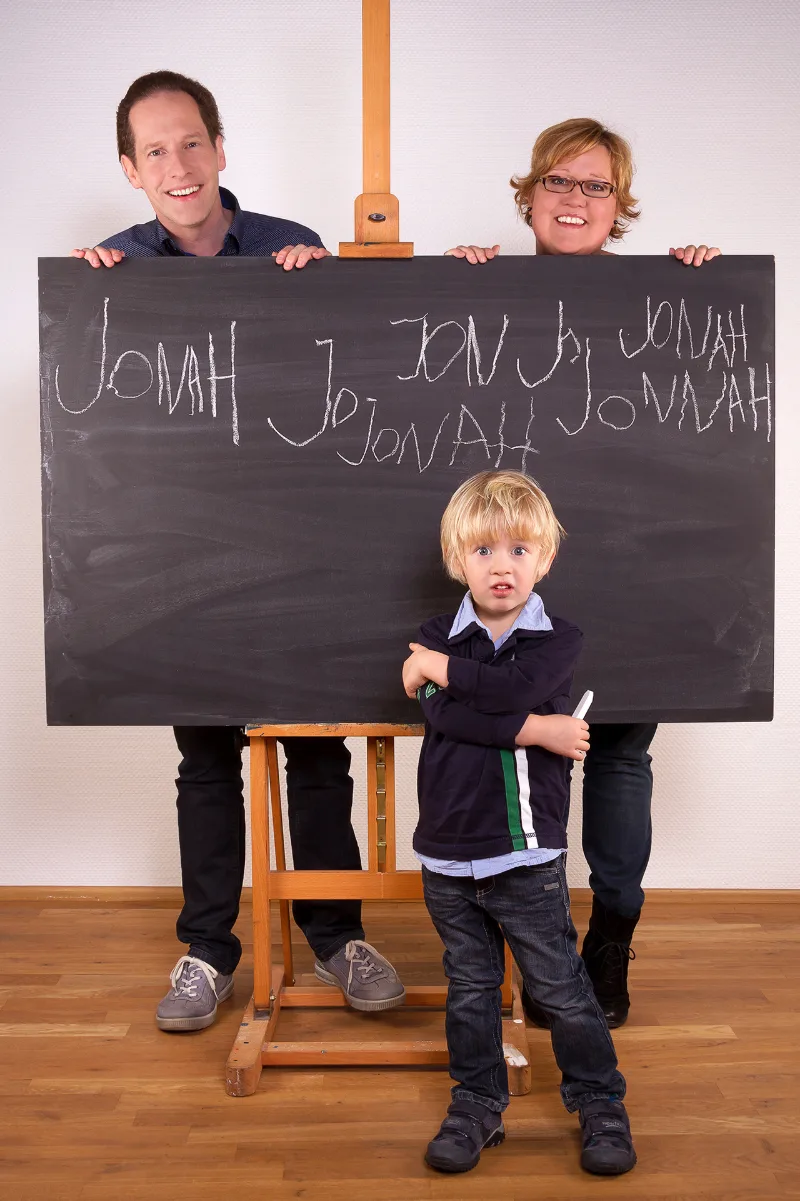 Parents holding a chalkboard on which a child's name is written many times; the boy stands in front holding a piece of chalk