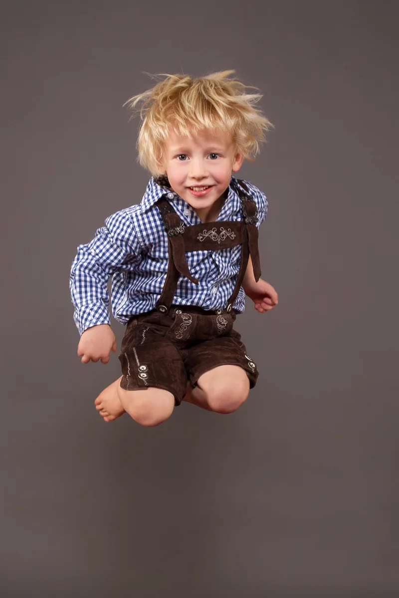 A blond boy in traditional dress jumping barefoot into the air, grey studio backdrop