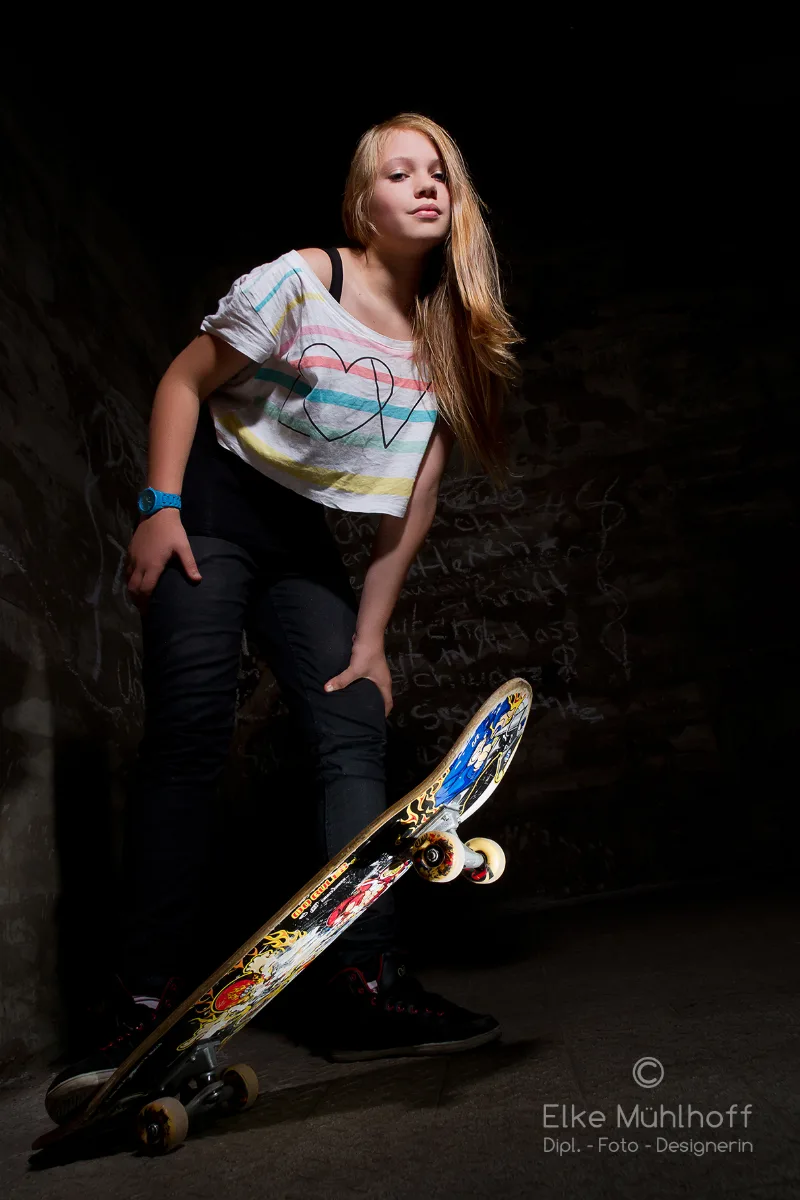 Mädchen mit Skateboard in einem dunklen Kellerraum, von oben angeleuchtet
