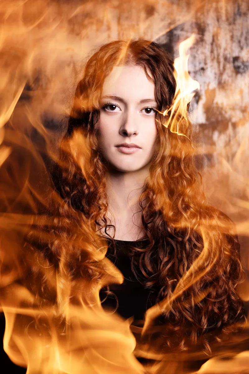 A creative portrait of a young woman with long curly hair, framed by real flames