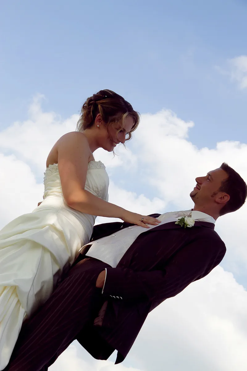 Bride and groom in wedding attire shot from below against a blue cloud-dotted sky