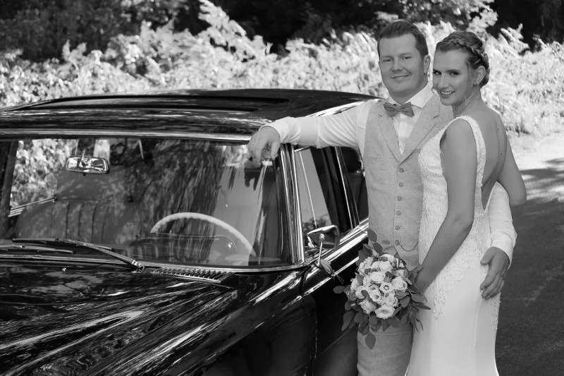 Bride and groom leaning casually against a black vintage car in warm sunlight (black and white)