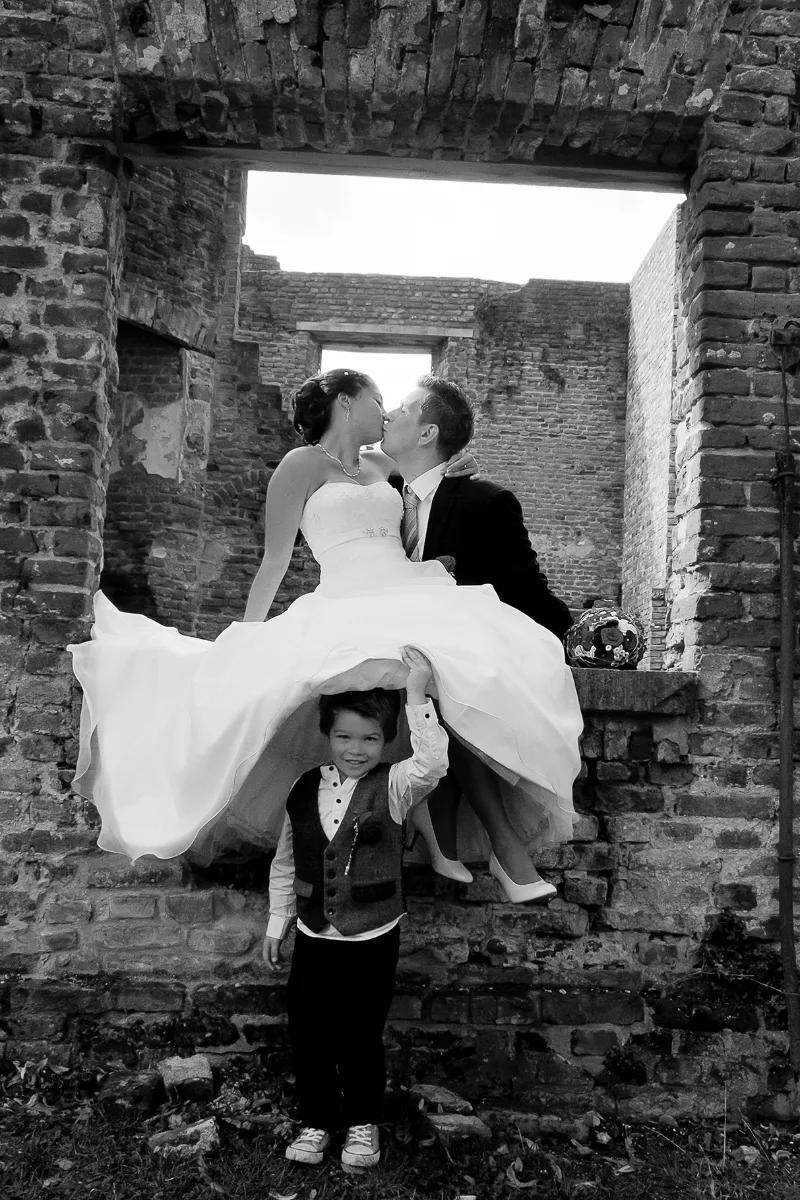 Bride and groom kissing in an old brick ruin, a flower child holding up the wedding dress