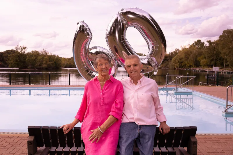 A couple in pink and rose on a wooden bench at the open-air pool, with silver helium balloons spelling 60 behind them