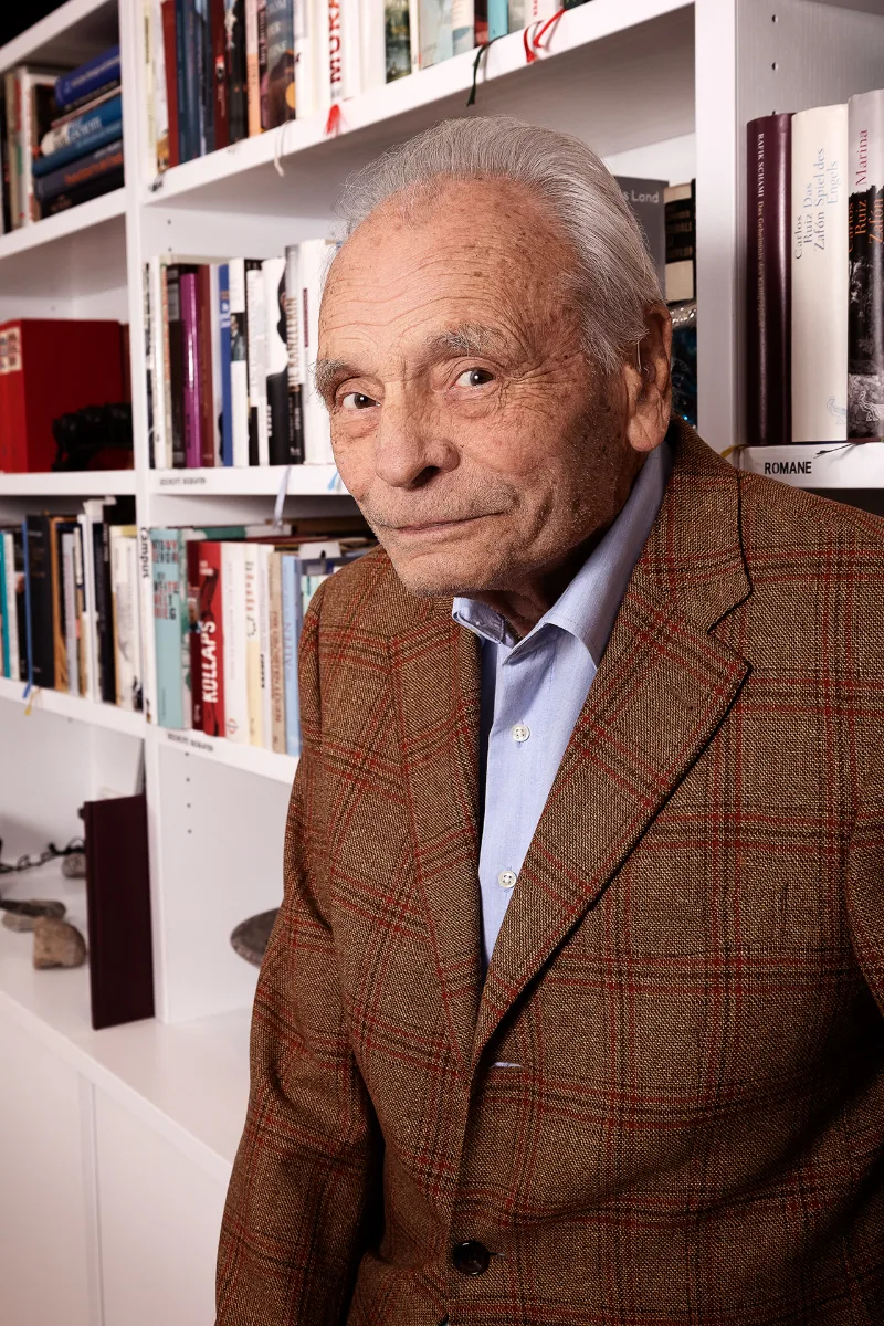 An older gentleman in a checked tweed jacket in front of a well-stocked white bookcase in daylight
