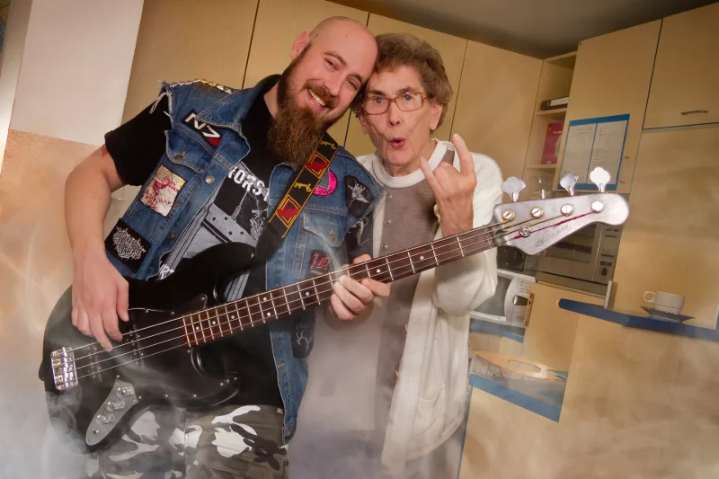 An elderly resident and a young heavy-metal fan with a bass guitar rocking out with a metal salute in a small kitchen, complete with stage smoke