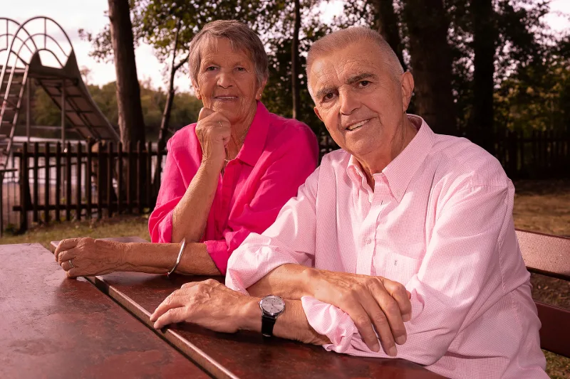 An older couple in pink and rose at a wooden table in the park, a lake in the background
